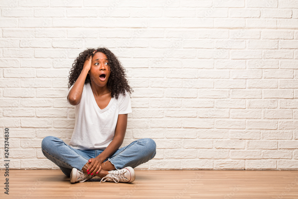 Young black woman sitting on a wooden floor worried and overwhelmed, forgetful, realize something, expression of shock at having made a mistake Young black woman sitting on a wooden floor worried and overwhelmed, forgetful, realize something, expression of shock at having made a mistake