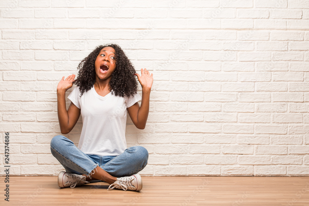 Young black woman sitting on a wooden floor screaming happy, surprised by an offer or a promotion, gaping, jumping and proud Young black woman sitting on a wooden floor screaming happy, surprised by an offer or a promotion, gaping, jumping and proud