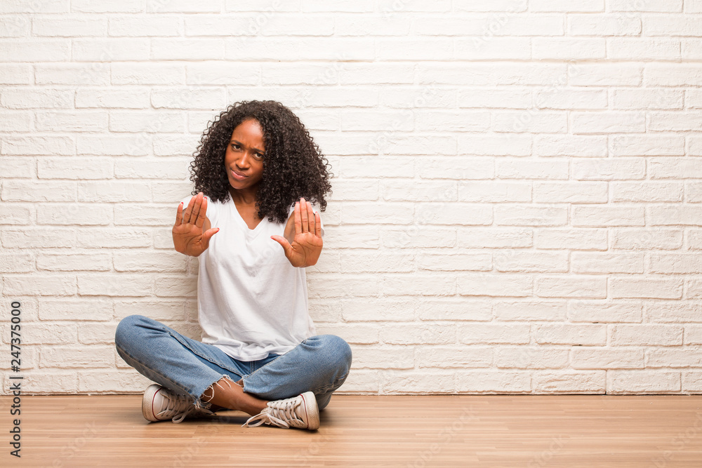 Young black woman sitting on wooden floor serious and determined, putting hand in front, stop gesture, deny concept Young black woman sitting on wooden floor serious and determined, putting hand in front, stop gesture, deny concept