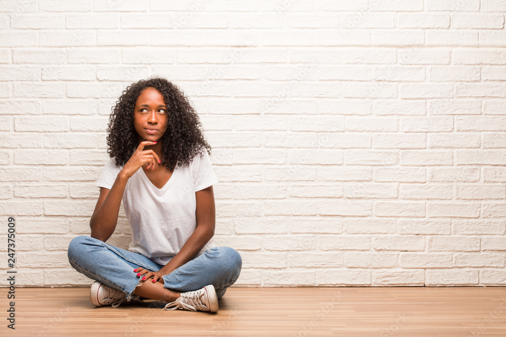 Young black woman sitting on wooden floor thinking and looking up, confused about an idea, would be trying to find a solution Young black woman sitting on wooden floor thinking and looking up, confused about an idea, would be trying to find a solution