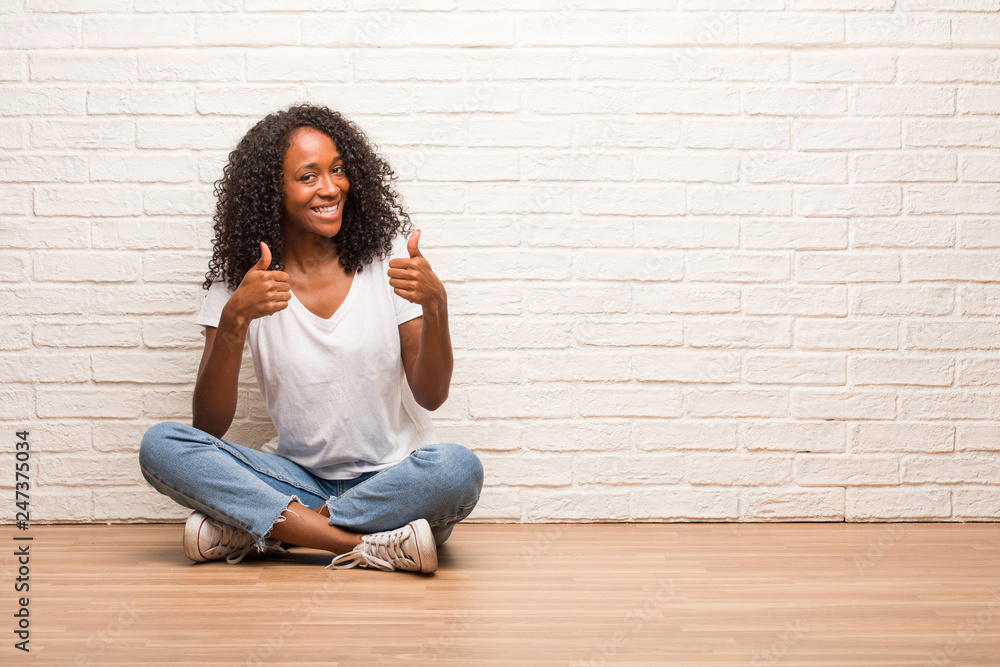 Young black woman sitting on wooden floor cheerful and excited, smiling and raising her thumb up, concept of success and approval, ok gesture Young black woman sitting on wooden floor cheerful and excited, smiling and raising her thumb up, concept of success and approval, ok gesture