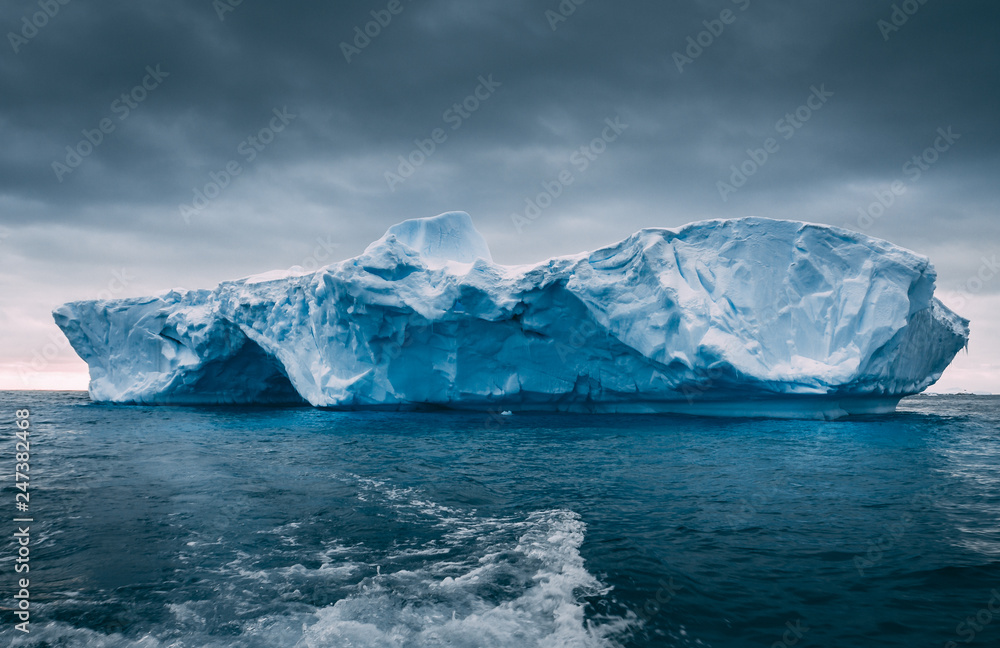 Big massive iceberg floating among the frozen ocean. Antarctica epic scenery in blue and grey tints. The dark cloudy sky over the South pole. The wild harsh environment. Mysterious winter landscape.