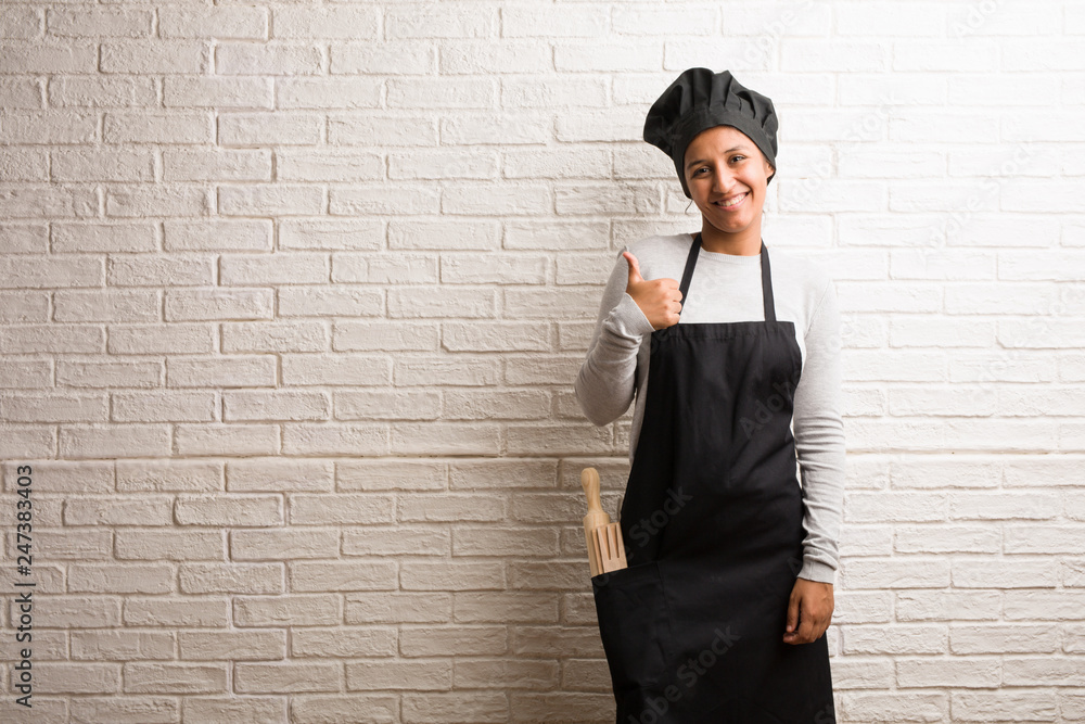 Young baker indian woman against a bricks wall cheerful and excited, smiling and raising her thumb up, concept of success and approval, ok gesture Young baker indian woman against a bricks wall cheerful and excited, smiling and raising her thumb up, concept of success and approval, ok gesture