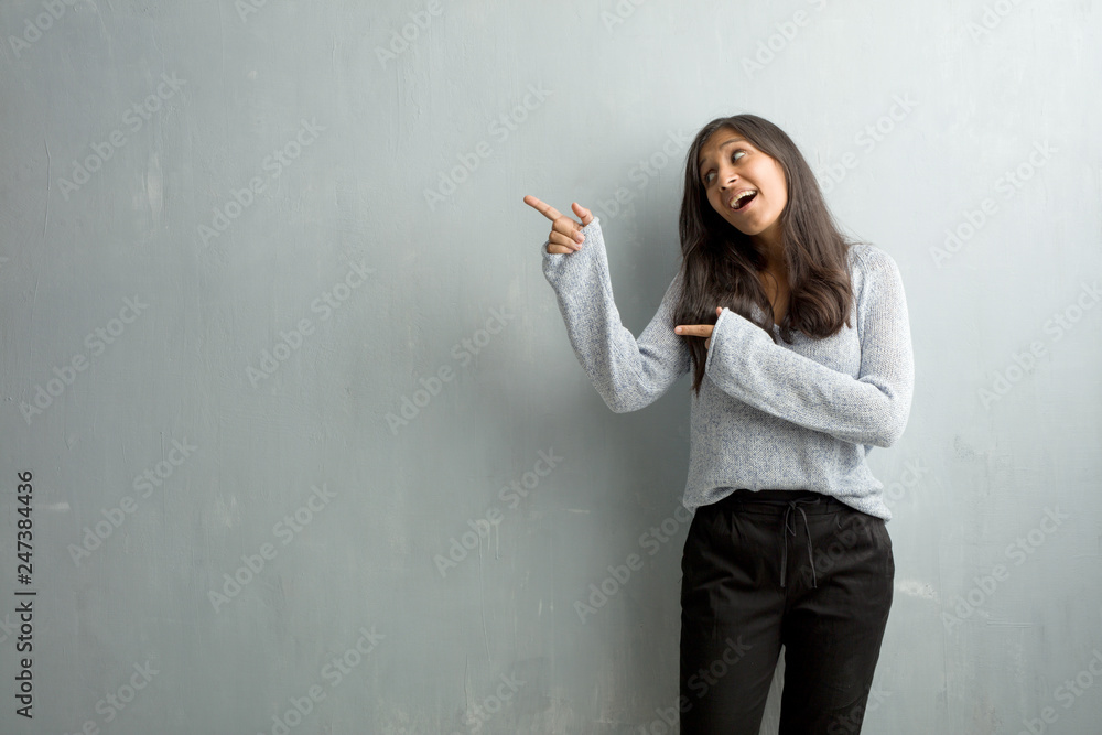 Young indian woman against a grunge wall pointing to the side, smiling surprised presenting something, natural and casual Young indian woman against a grunge wall pointing to the side, smiling surprised presenting something, natural and casual