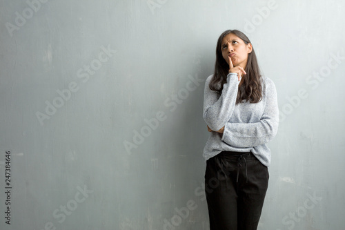 Young indian woman against a grunge wall thinking and looking up, confused about an idea, would be trying to find a solution