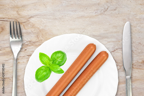 Tasty boiled frankfurter sausages decorated with basil leaves on a plate with fork and knife on on the marble countertop photo