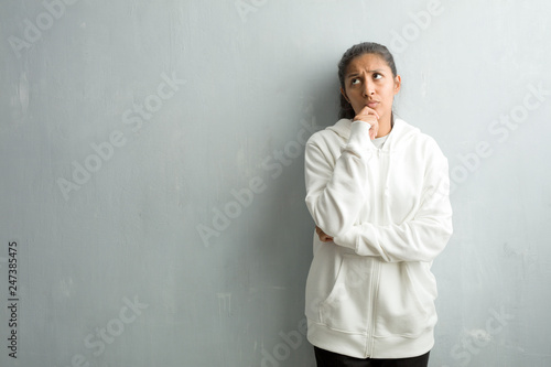 Young sporty indian woman against a gym wall doubting and confused, thinking of an idea or worried about something