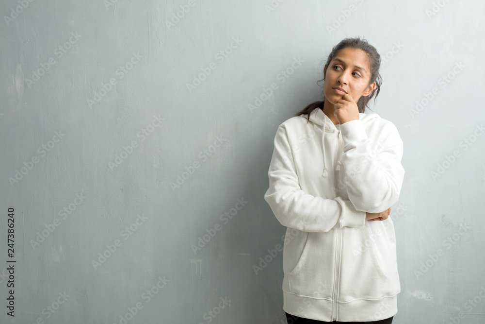 Young sporty indian woman against a gym wall thinking and looking up, confused about an idea, would be trying to find a solution Young sporty indian woman against a gym wall thinking and looking up, confused about an idea, would be trying to find a solution
