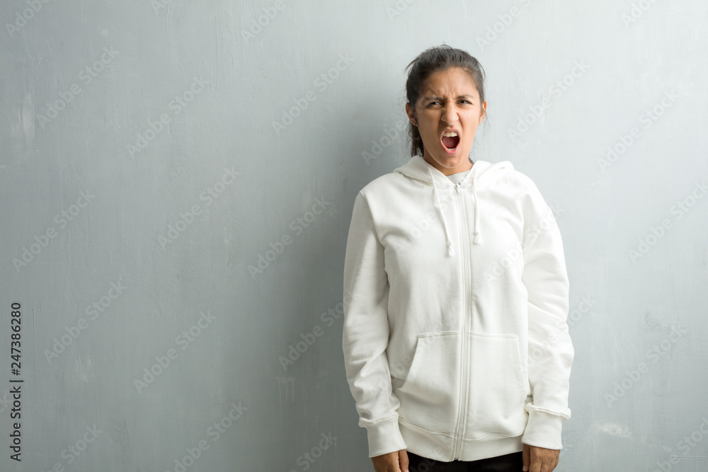 Young sporty indian woman against a gym wall very angry and upset, very tense, screaming furious, negative and crazy