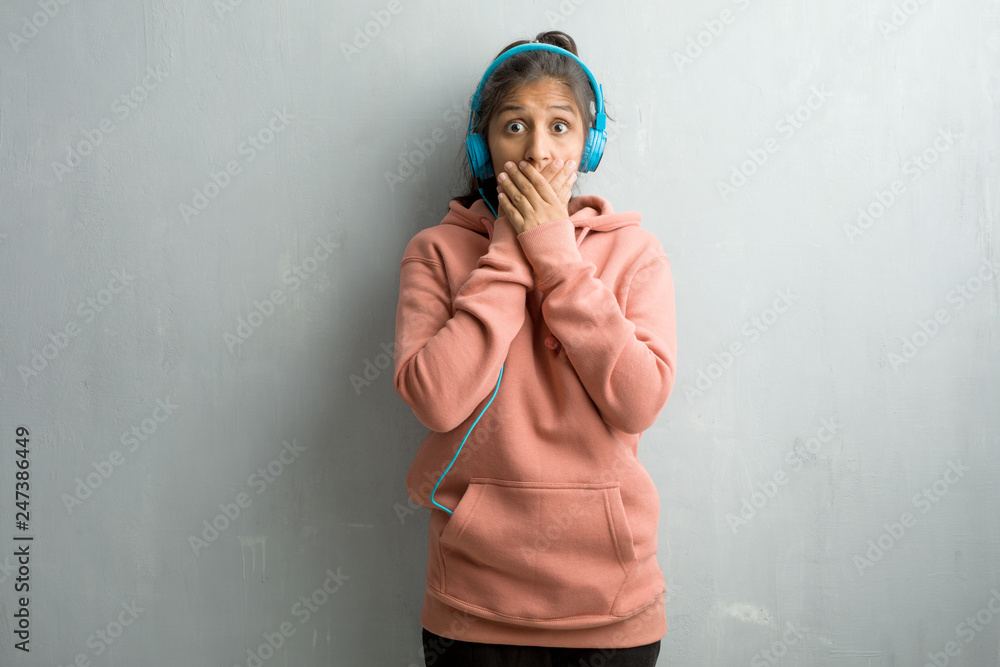 Fototapeta premium Young sporty indian woman against a wall covering mouth, symbol of silence and repression, trying not to say anything. Listening music with headphones.