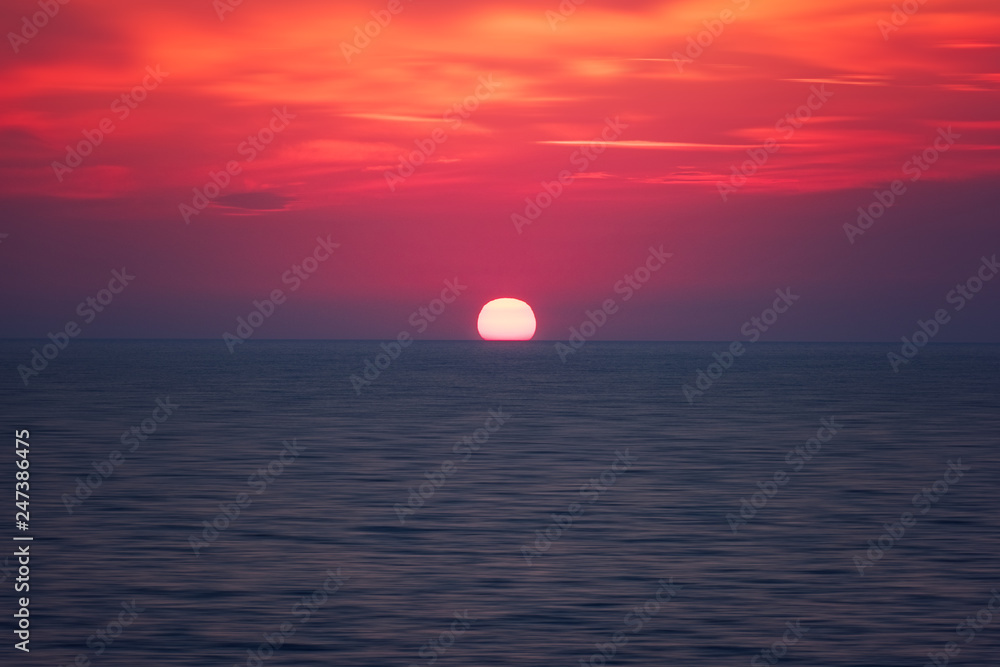 Scenic sunset seascape with sea, sun and amazing scarlet cloudy sky, outdoor travel background, motion blur