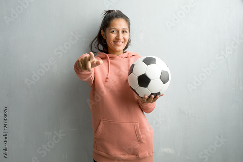 Young sporty indian woman against a wall cheerful and smiling pointing to the front. Holding a soccer ball to play a game. © Asier