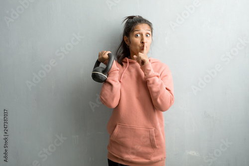 Young sporty indian woman against a wall keeping a secret or asking for silence, serious face, obedience concept. Holding a dumbbell with hand.