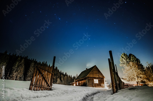 Rural wooden cottage in the mountains with windows glowing in the dark at winter night. Wooden hut on hill moutains with old fence with blue starry sky above