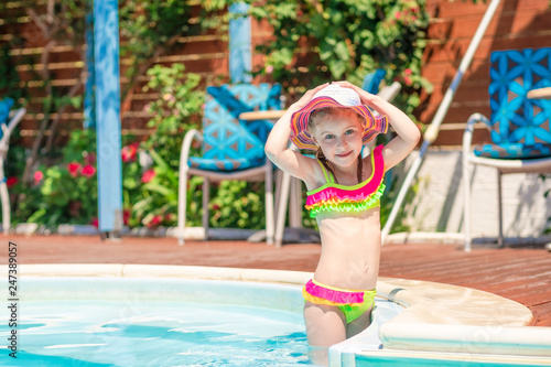 Little smiling blond girl in a bright swimsuit holding her hat standing in the blue pool