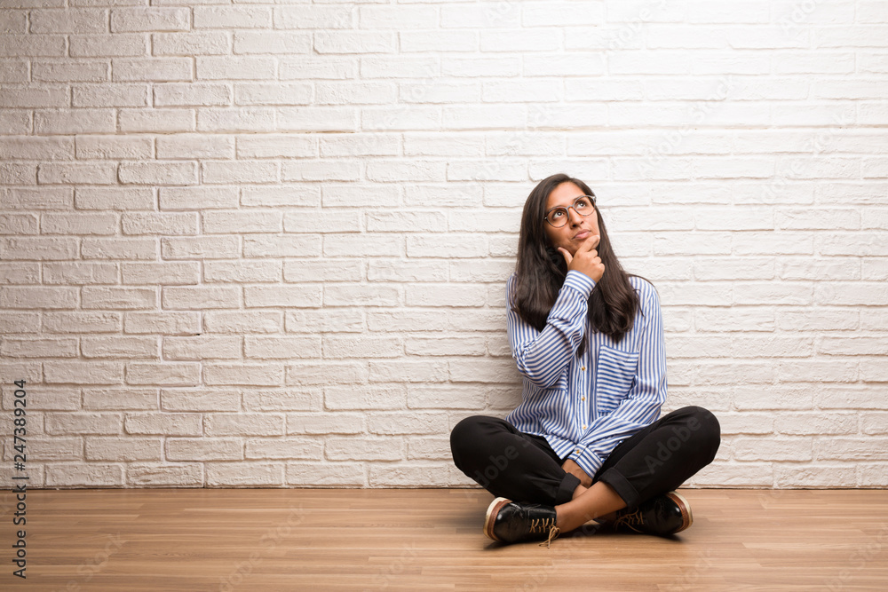 Young indian woman sit against a brick wall doubting and confused, thinking of an idea or worried about something Young indian woman sit against a brick wall doubting and confused, thinking of an idea or worried about something
