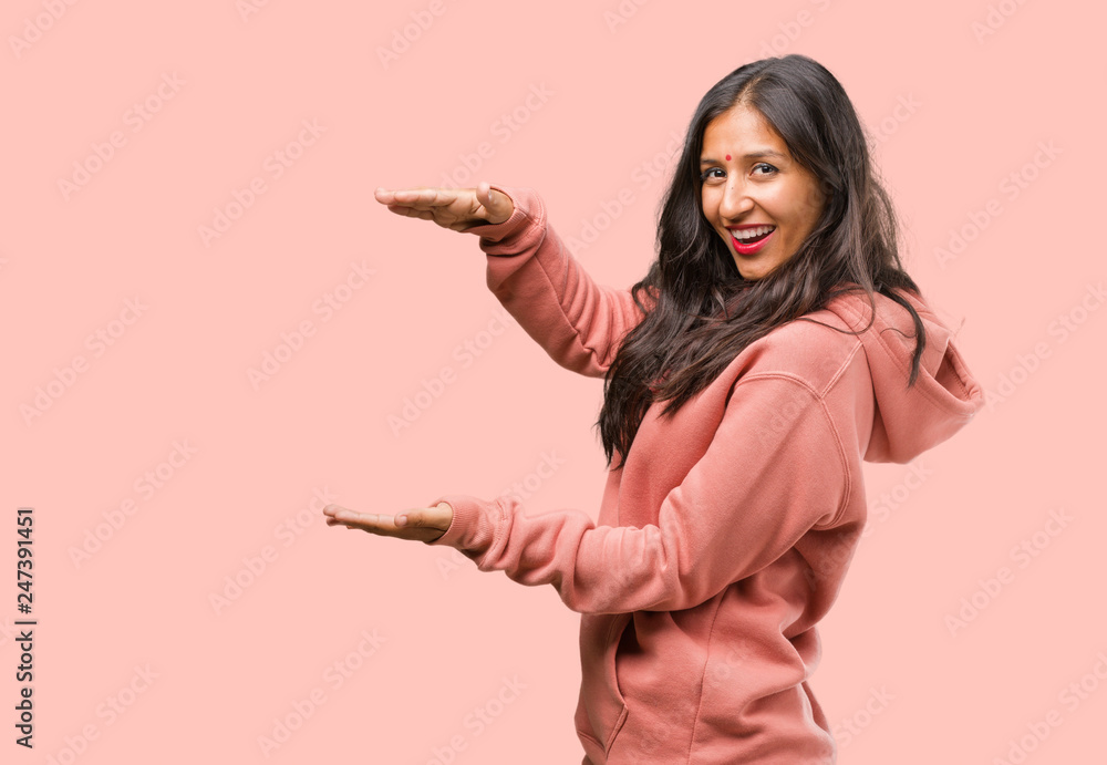 Portrait of fitness young indian woman holding something with hands, showing a product, smiling and cheerful, offering an imaginary object Portrait of fitness young indian woman holding something with hands, showing a product, smiling and cheerful, offering an imaginary object