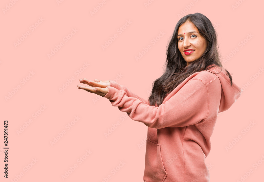 Portrait of fitness young indian woman holding something with hands, showing a product, smiling and cheerful, offering an imaginary object Portrait of fitness young indian woman holding something with hands, showing a product, smiling and cheerful, offering an imaginary object