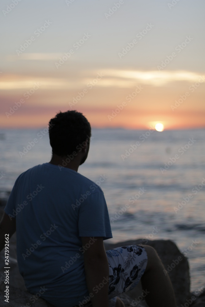 Young man sitting alone on the seashore during sunset. Warm, pleasant, summer evening on the Croatian coast. Calm end of the day on the shore. Vacation, rest in Croatia. Dusk, twilight, Adriatic Sea Young man sitting alone on the seashore during sunset. Warm, pleasant, summer evening on the Croatian coast. Calm end of the day on the shore. Vacation, rest in Croatia. Dusk, twilight, Adriatic Sea
