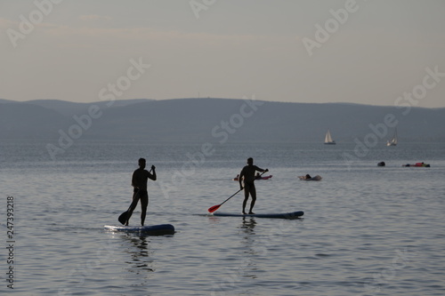 Two men floating on boards. Stand up paddle on Lake Balaton. Beautiful, warm, sunny, calm summer day. Mountains, sailboats, blue sky visible in the background. Active rest during the holidays. © Mateusz