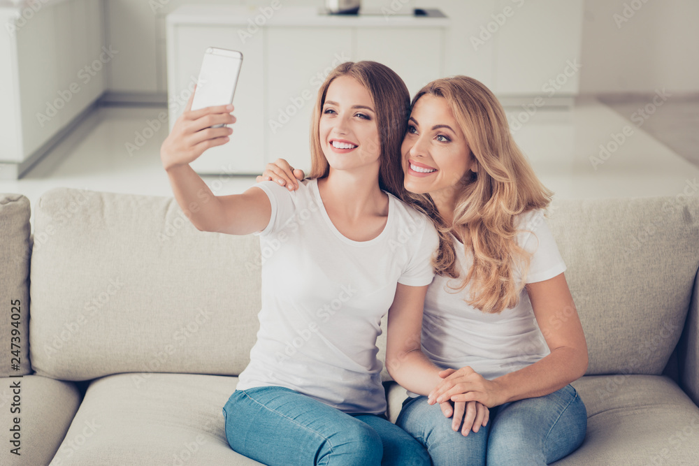 Two nice cute attractive charming lovely winsome lovable fascinating tender cheerful ladies sitting on divan using gadget making taking selfie in light white interior room indoors Two nice cute attractive charming lovely winsome lovable fascinating tender cheerful ladies sitting on divan using gadget making taking selfie in light white interior room indoors