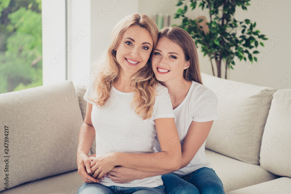 Close up photo of two people pretty mum mommy and teen daughter holding each other hands arms piggy back lovely beaming toothy smiling wear white t-shirts jeans sit on comfy sofa couch Close up photo of two people pretty mum mommy and teen daughter holding each other hands arms piggy back lovely beaming toothy smiling wear white t-shirts jeans sit on comfy sofa couch