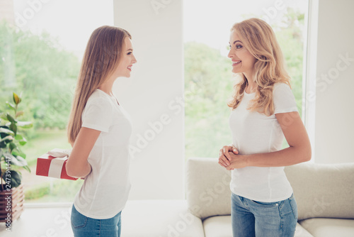 Close up side profile view photo cheer toothy smiling two people mum and teen daughter holding hands arms behind back with gift box surprise wear white t-shirts jeans in bright kitchen