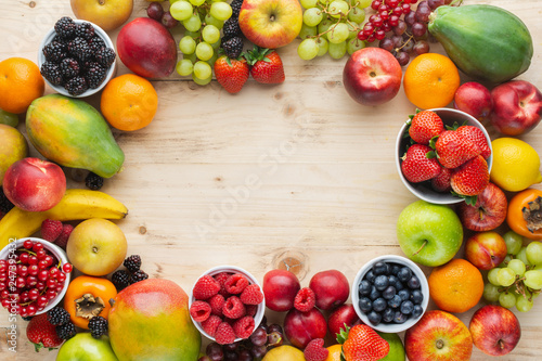 Rainbow fruits background frame  strawberries raspberries oranges plums apples kiwis grapes blueberries mango persimmon on light wooden table  top view  copy space for text  selective focus