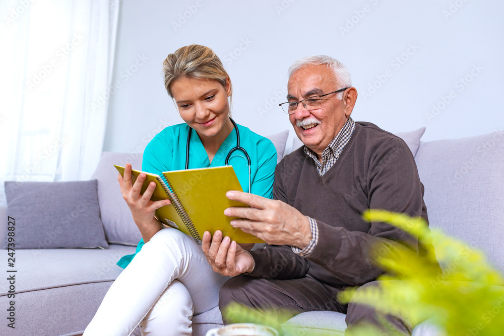 Woman reading a book while sitting with happy grandfather. Happy elder man sitting on white sofa and listening to nurse reading a book out loud Woman reading a book while sitting with happy grandfather. Happy elder man sitting on white sofa and listening to nurse reading a book out loud