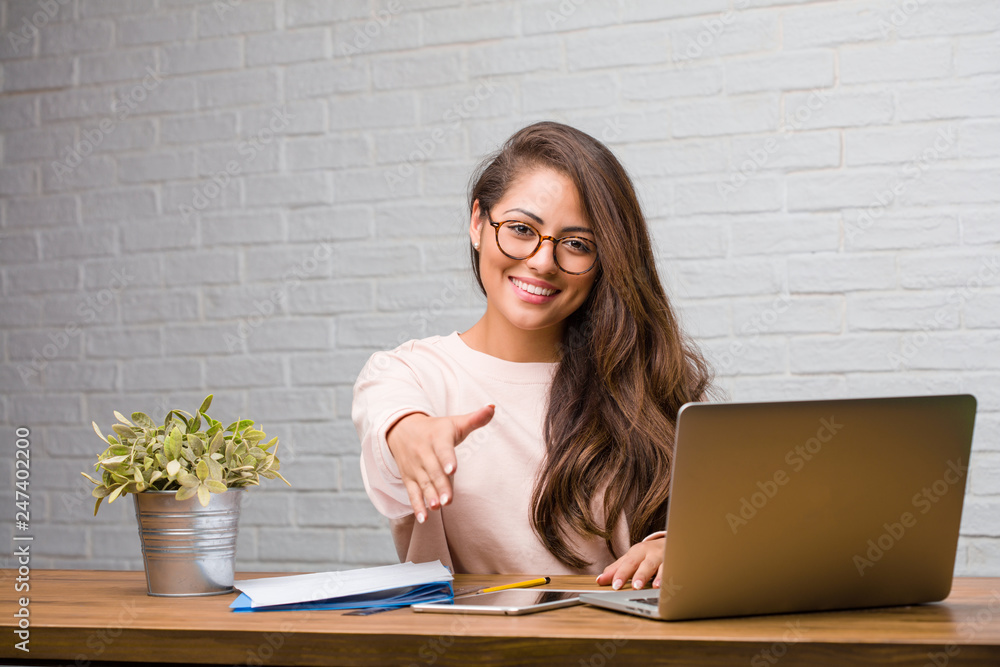 Portrait of young student latin woman sitting on her desk reaching out to greet someone or gesturing to help, happy and excited Portrait of young student latin woman sitting on her desk reaching out to greet someone or gesturing to help, happy and excited