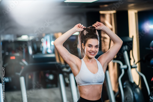 Ready to workout! Beautiful smiling girl in sportswear tying her hair while standing in the gym, looking at camera.