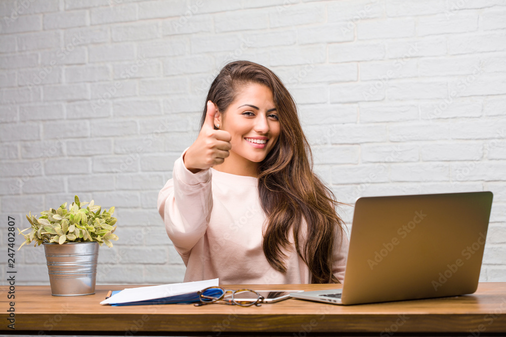 Portrait of young student latin woman sitting on her desk cheerful and excited, smiling and raising her thumb up, concept of success and approval, ok gesture Portrait of young student latin woman sitting on her desk cheerful and excited, smiling and raising her thumb up, concept of success and approval, ok gesture