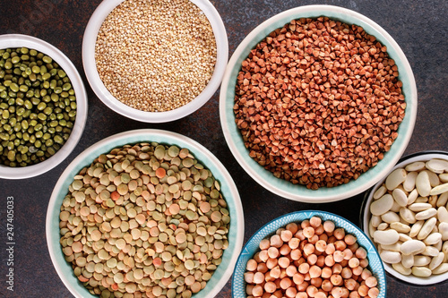 Top view on bowls with various raw grains and seeds on a textured dark background.