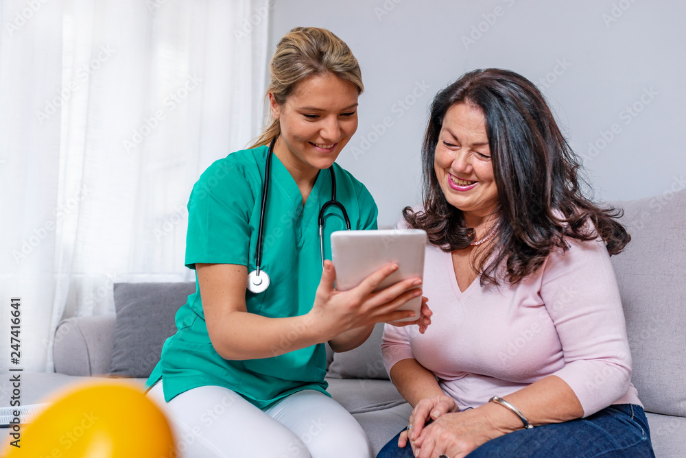Elderly woman sitting in the living room with home health care nurse and consulting. Middle age caregiver holding in hand digital tablet and giving advice to her patient. Elderly woman sitting in the living room with home health care nurse and consulting. Middle age caregiver holding in hand digital tablet and giving advice to her patient.