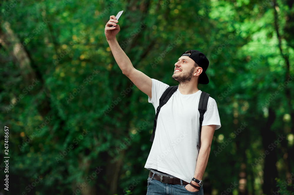 Smiling male taking a selfie against green trees. Man traveler in summer clothing standing outdoors taking selfie with her mobile phone