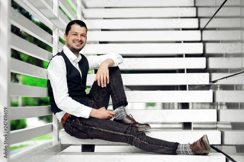 Image of young male in casual outfit, laughing while holding a phone in hands outdoors.