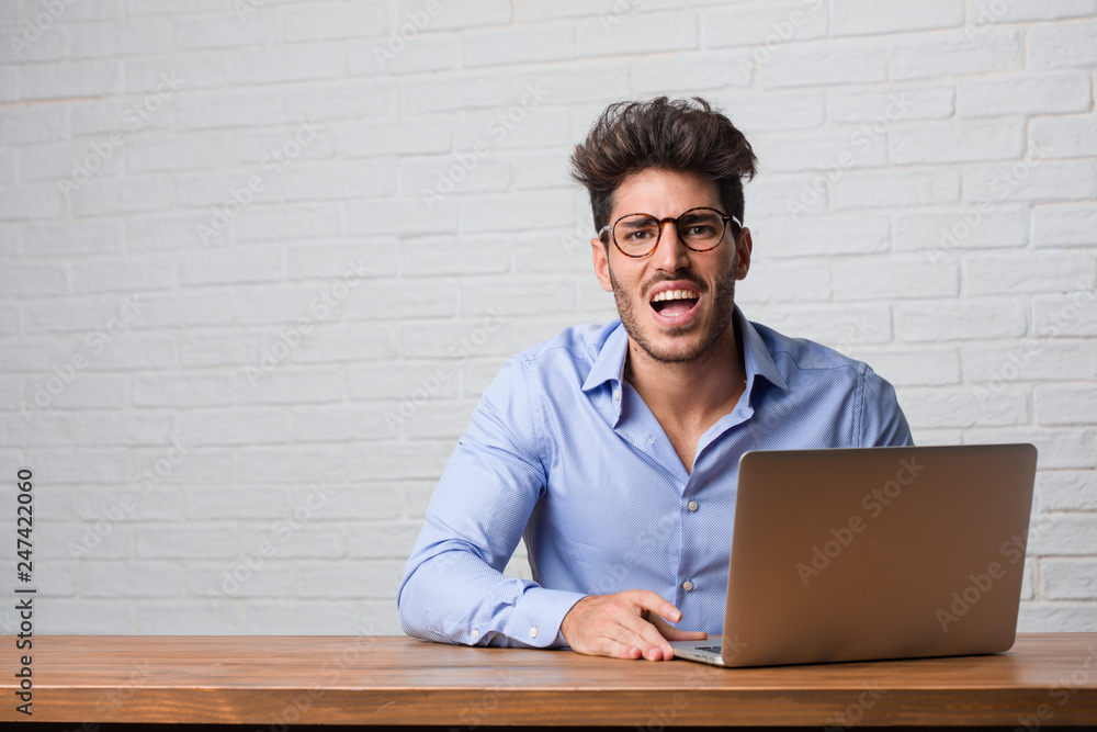 Young business man sitting and working on a laptop screaming angry, expression of madness and mental instability, open mouth and half-opened eyes, madness concept Young business man sitting and working on a laptop screaming angry, expression of madness and mental instability, open mouth and half-opened eyes, madness concept