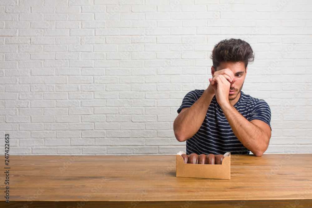 Young handsome and natural man sitting on a table looking through a gap, hiding and squinting. Eating chocolate doughnuts. Young handsome and natural man sitting on a table looking through a gap, hiding and squinting. Eating chocolate doughnuts.