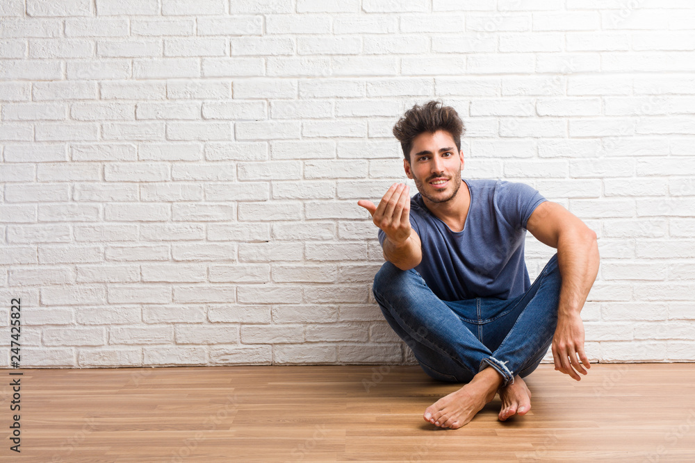 Young natural man sit on a wooden floor inviting to come, confident and smiling making a gesture with hand, being positive and friendly Young natural man sit on a wooden floor inviting to come, confident and smiling making a gesture with hand, being positive and friendly