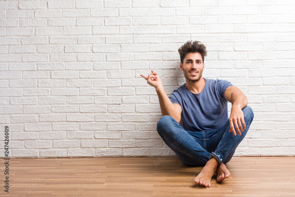Young natural man sit on a wooden floor pointing to the side, smiling surprised presenting something, natural and casual Young natural man sit on a wooden floor pointing to the side, smiling surprised presenting something, natural and casual