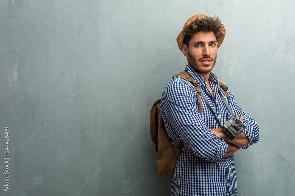 Young handsome traveler man wearing a straw hat, a backpack and a photo camera crossing his arms, smiling and happy, being confident and friendly