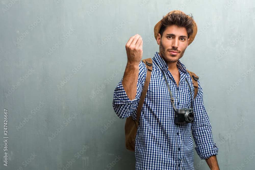Young handsome traveler man wearing a straw hat, a backpack and a photo camera doing a typical italian gesture, smiling and looking straight ahead, symbol or expression with hand, very natural