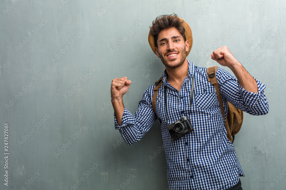 Young handsome traveler man wearing a straw hat, a backpack and a photo camera very happy and excited, raising arms, celebrating a victory or success, winning the lottery