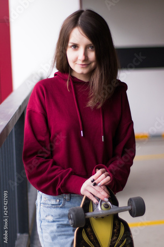 A girl with big eyes and a longboard in her arms, in a burgundy hoodie