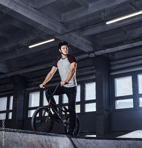 Full body portrait of a young man in protective helmet with his bike standing in a skatepark indoors