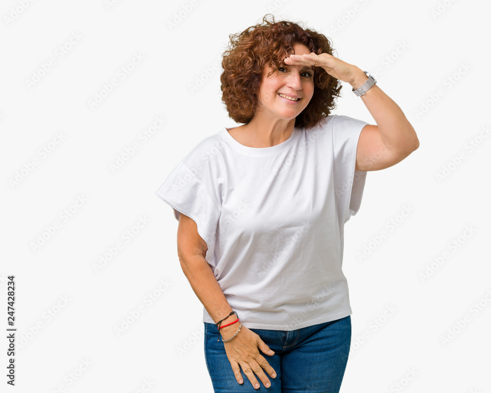 Beautiful middle ager senior woman wearing white t-shirt over isolated background very happy and smiling looking far away with hand over head. Searching concept. Beautiful middle ager senior woman wearing white t-shirt over isolated background very happy and smiling looking far away with hand over head. Searching concept.
