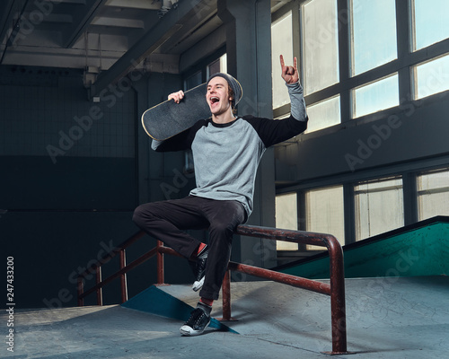 Cheerful young man wearing a shirt and hat holding his board, sitting on a grind rail in skatepark indoors.
