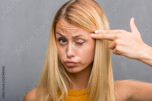 Face closeup, portrait of a natural young blonde woman making a suicide gesture, feeling sad and scared forming a gun with fingers