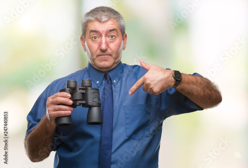 Handsome senior man looking through binoculars over isolated background with surprise face pointing finger to himself