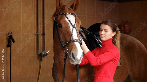 Beautiful girl with her horse in stable. Young woman adjusting her horse bridle before a ride. People and animals concept. Equine business. © DenisProduction.com